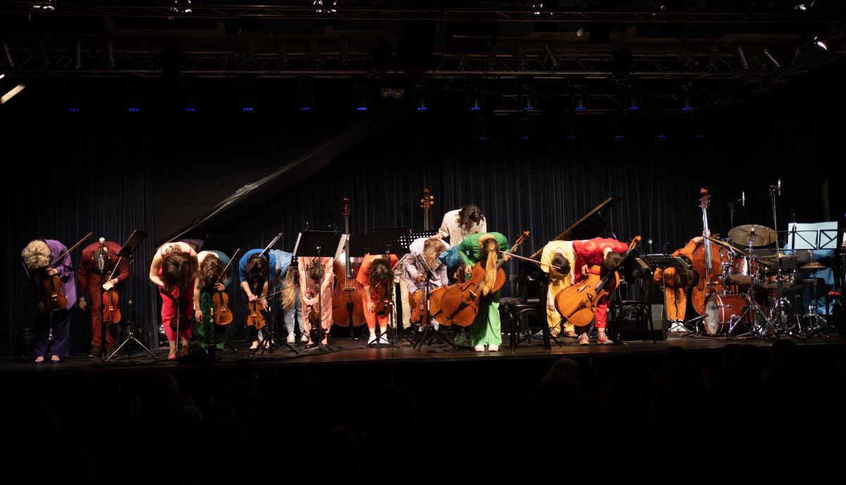 Bunt gekleidete Musiker:innen mit Instrumenten verneigen sich auf einer Bühne